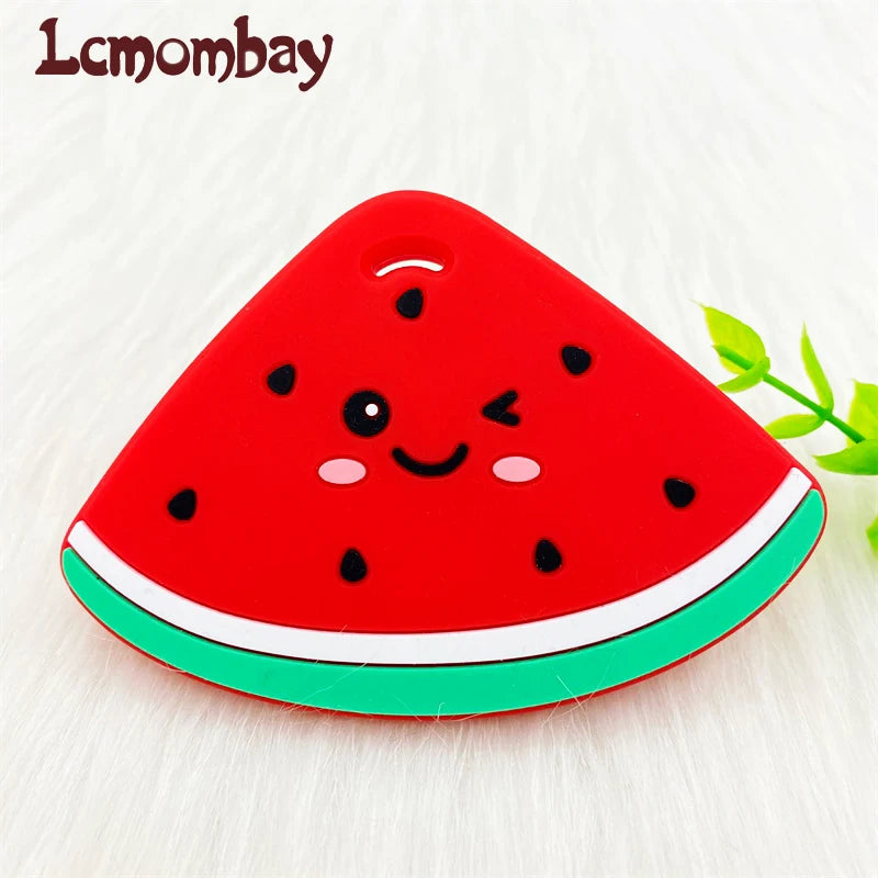 A bright red watermelon silicone teether pendant with black seeds and a green rind, resting on a white surface.