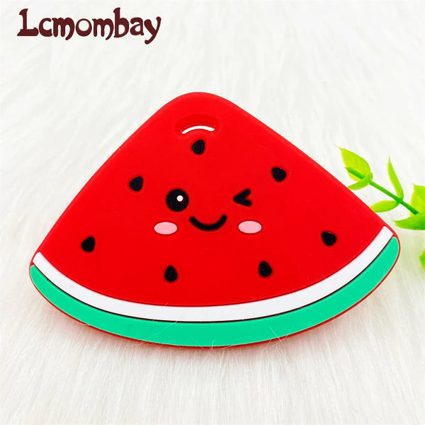 A bright red watermelon silicone teether pendant with black seeds and a green rind, resting on a white surface.