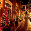A row of Christmas trees decorated with warm string lights lines a brick pathway at night.