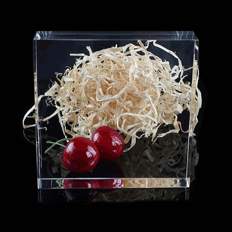 A clear acrylic sheet contains a pile of light brown shredded material and a red cherry fruit.