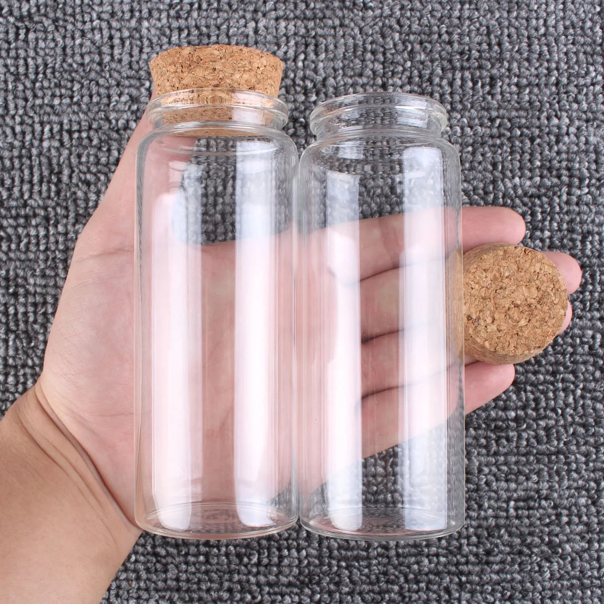 Una mano sostiene dos frascos de vidrio transparente con tapones de corcho contra un fondo gris texturizado.