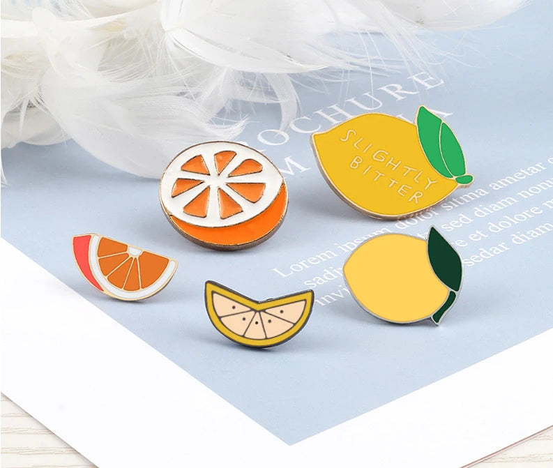 A collection of enamel fruit brooches featuring orange, lemon, and citrus designs on a light blue background.