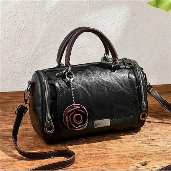 A black leather crossbody shoulder bag with a brown strap and a pink floral pendant resting on a wooden surface.