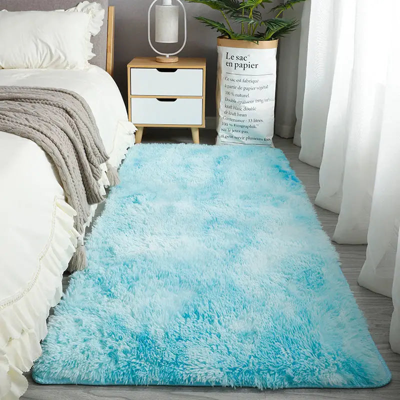 A bright turquoise fluffy rug placed in front of a white bed with light gray bedding and a wooden side table.