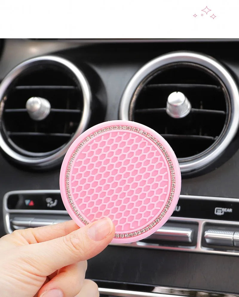 A hand holds a pink crystal rhinestone non-slip cup mat for cars, positioned in front of car air vents.