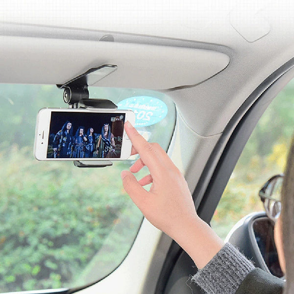 A hand holds a smartphone mounted on a car sun visor phone holder inside a vehicle.