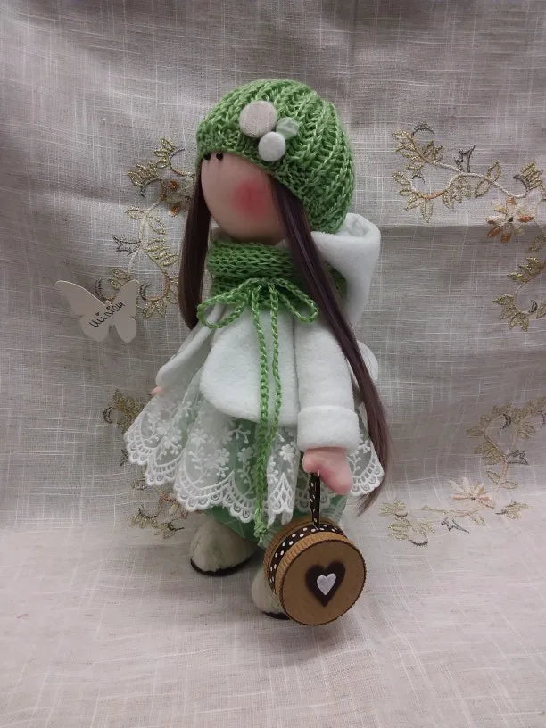 A handmade doll with long dark hair wearing a green knitted hat and white clothing.