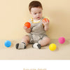 A toddler is holding an orange ball in front of a light yellow background with various colored ocean balls scattered on the s