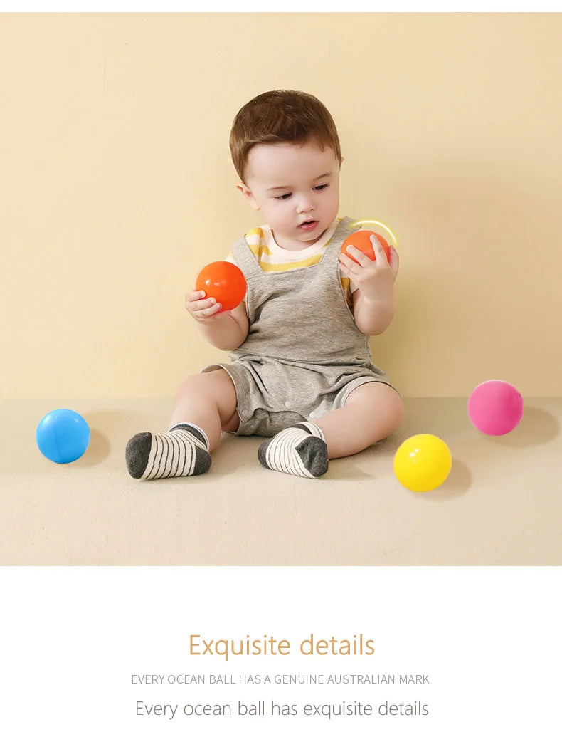 A toddler is holding an orange ball in front of a light yellow background with various colored ocean balls scattered on the s