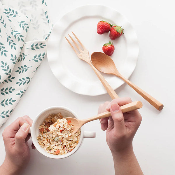 A set of natural beech wood cutlery, including a fork, spoon, and a wooden spoon, placed on a white plate with strawberries.