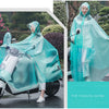 A woman wearing a transparent, iridescent waterproof motorcycle rain poncho with a hood is riding a scooter outdoors.
