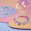 A colorful bracelet made of polymer clay beads with letters is displayed on a pink and purple background.