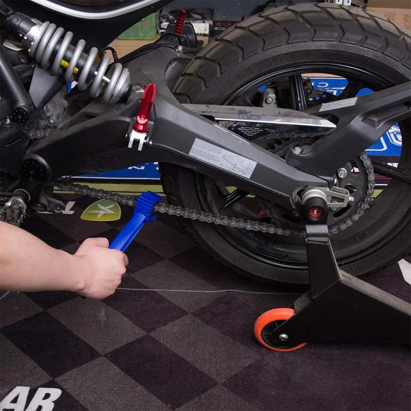 A hand is using a blue chain cleaning brush on the drive chain of a motorcycle or bicycle.