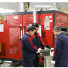 Three workers inspecting a red industrial machine with plastic buckets and lids.