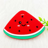 A bright red watermelon silicone teether pendant with black seeds and a green rim resting on white fur.