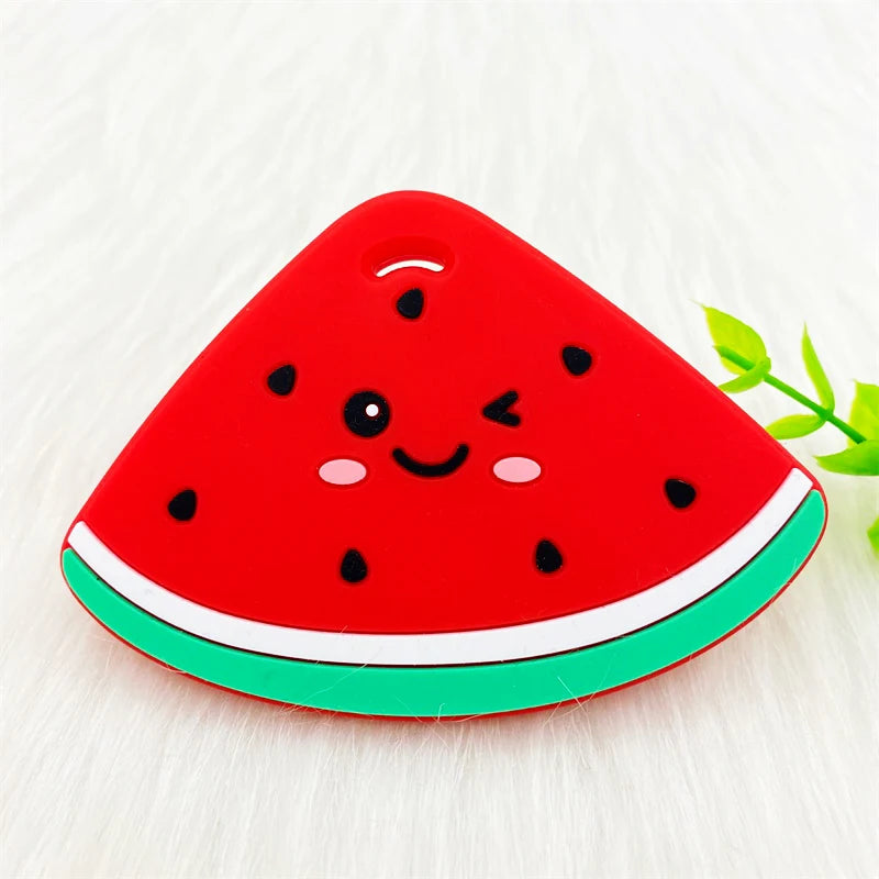A bright red watermelon silicone teether pendant with black seeds and a green rim resting on white fur.