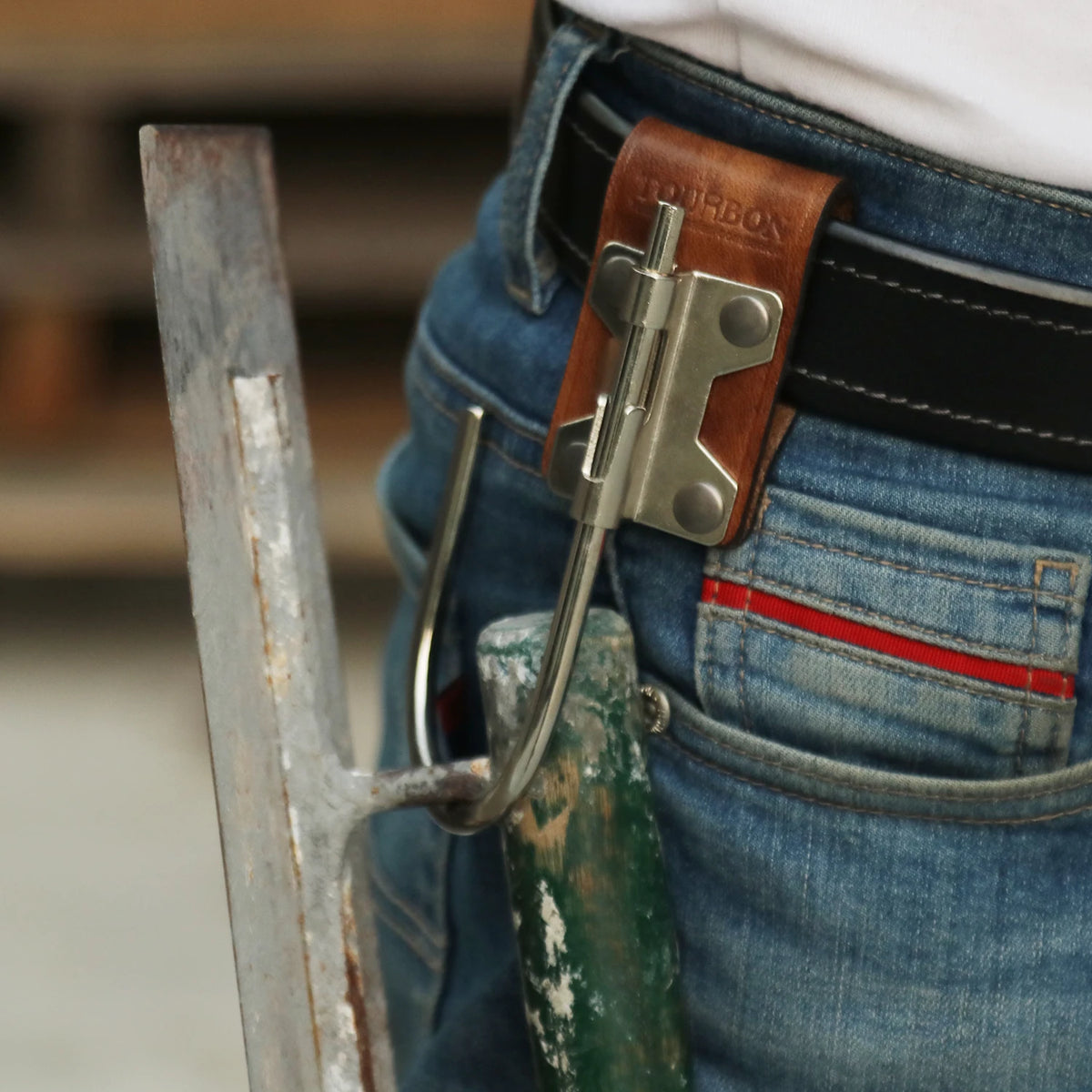 A close-up of a brown leather tool holster hook with a steel J-hook attached to denim jeans.