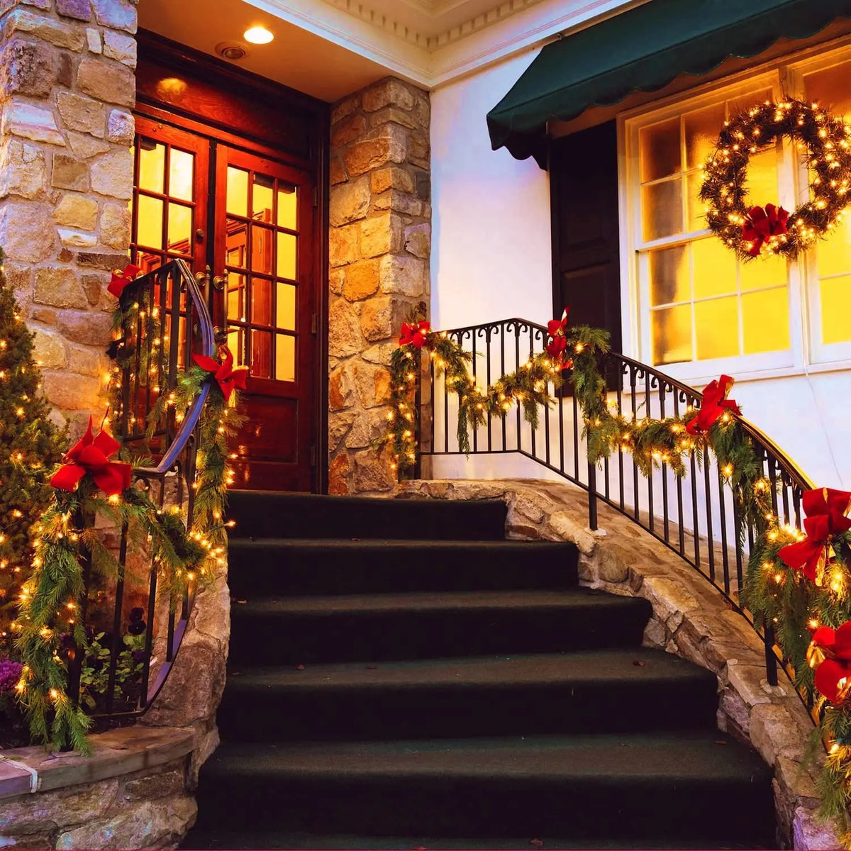 A stone staircase and railing decorated with warm string lights and red flowers leading to a wooden door.