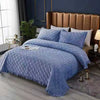 A blue quilted velvet bedspread with a ruffled edge is displayed on a bed against a brown upholstered headboard.