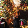 A young girl stands in a dimly lit room surrounded by a large decorated Christmas tree with warm string lights.