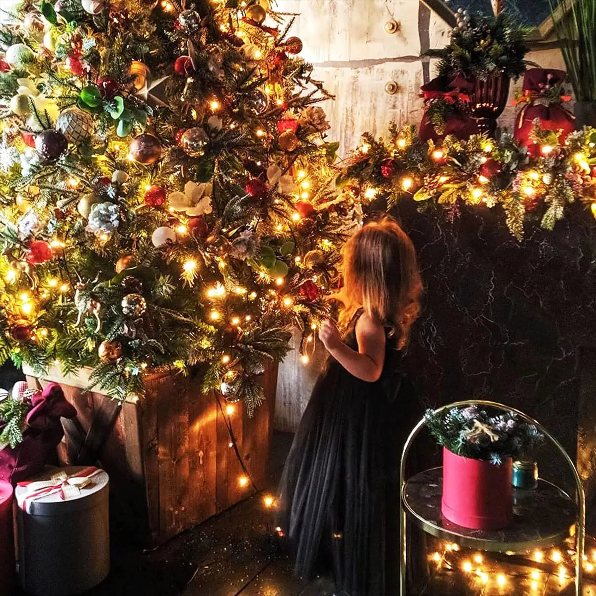 A young girl stands in a dimly lit room surrounded by a large decorated Christmas tree with warm string lights.