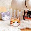 An embroidered orange and white cat patch resting on a beige fabric next to a white picket fence and a fluffy white animal.