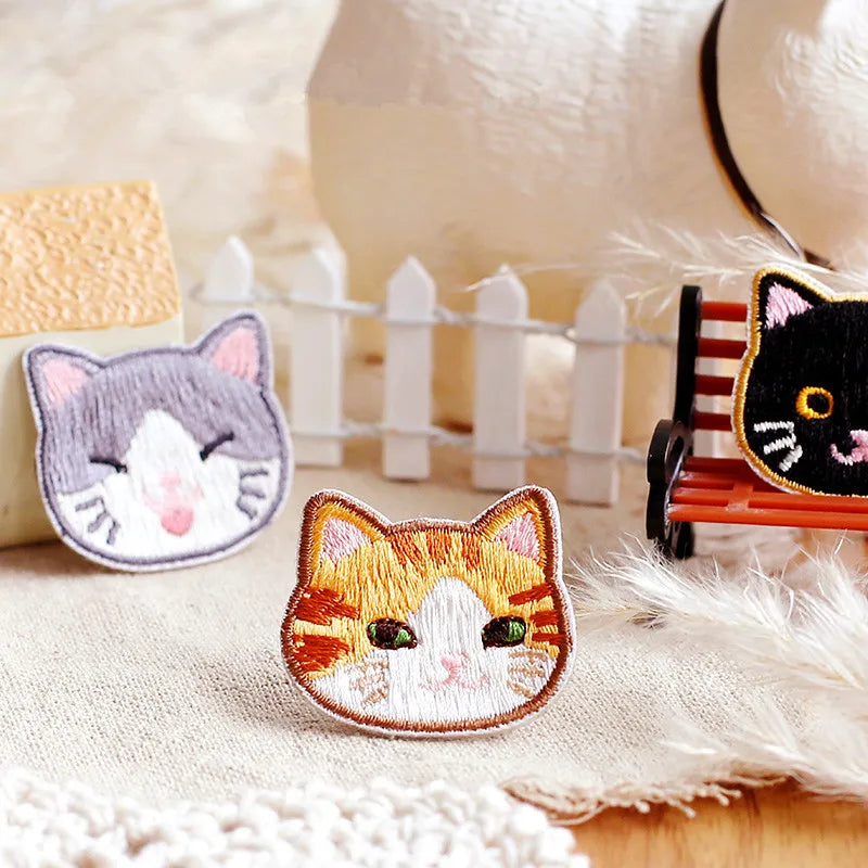 An embroidered orange and white cat patch resting on a beige fabric next to a white picket fence and a fluffy white animal.