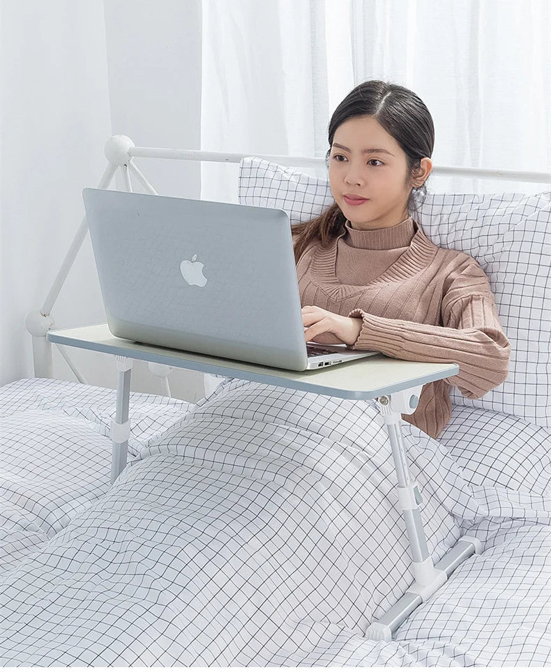 A woman is using an adjustable foldable laptop desk placed on a bed, working on a silver laptop.