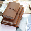 A stack of brown leather refillable journals resting on a wooden surface next to paper and a grid.