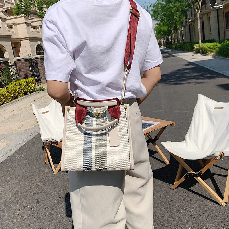 A person wearing a white shirt and light pants holds a beige canvas crossbody tote bag with red strap.