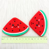 A bright red watermelon silicone teether pendant with green trim resting on a white, fluffy surface.