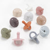 A collection of various shades of silicone baby pacifiers and soothers arranged on a white background.