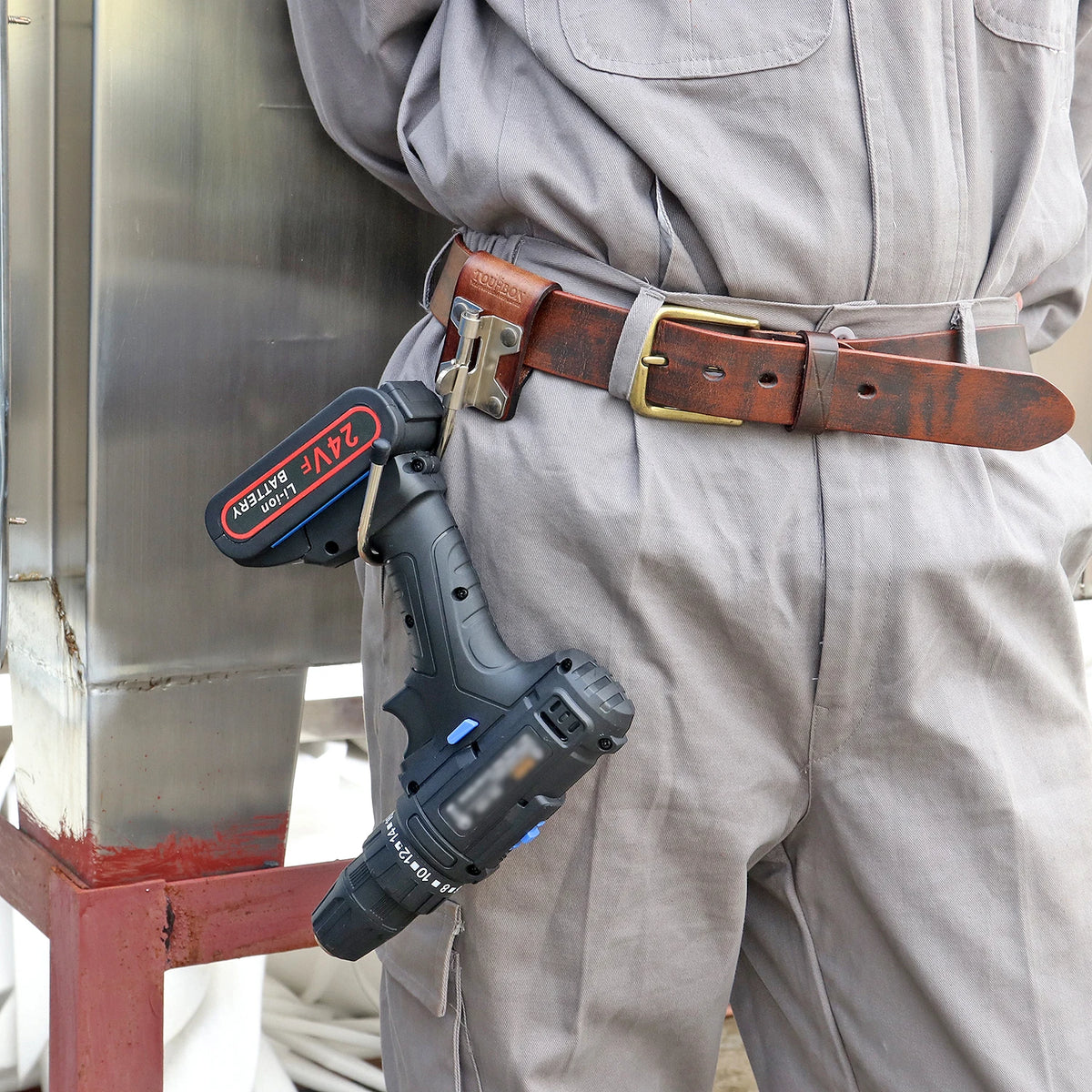A brown leather tool holster featuring a steel J-hook attached to a belt, worn on light gray pants.