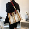 A person holds a beige and brown striped canvas crossbody tote bag with a leather strap against a white background.