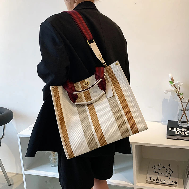 A person holds a beige and brown striped canvas crossbody tote bag with a leather strap against a white background.