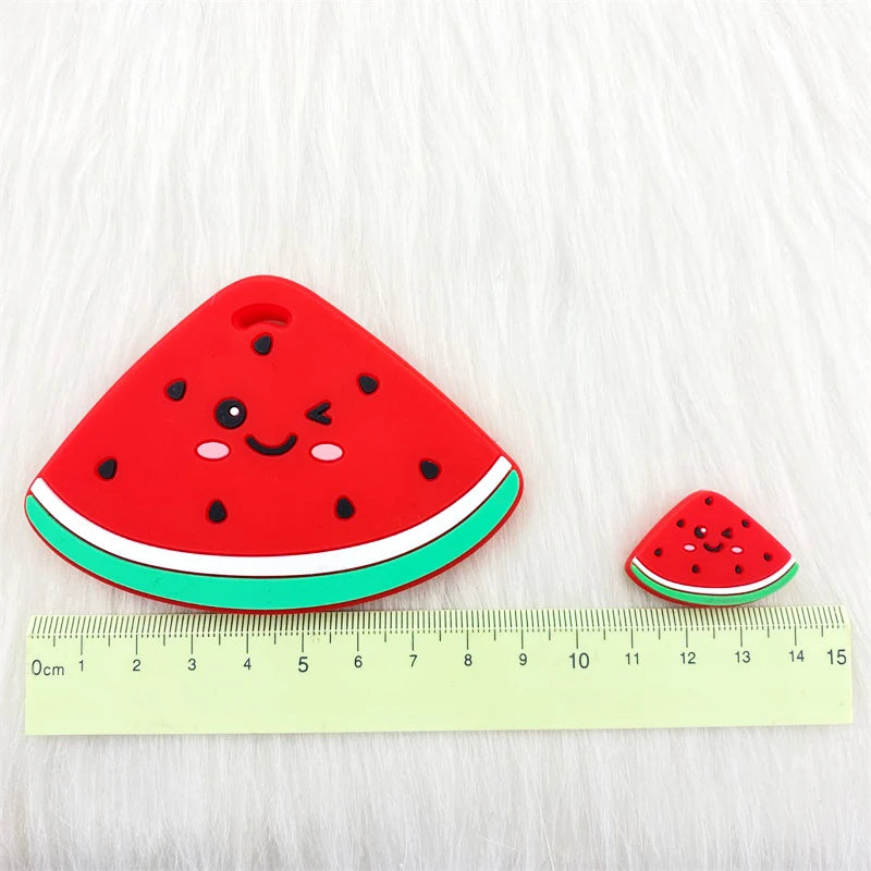 A red watermelon silicone teether pendant resting on a yellow ruler against a white, fluffy background.