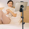 A woman is reading a book while using an adjustable aluminum tripod stand to hold a tablet.