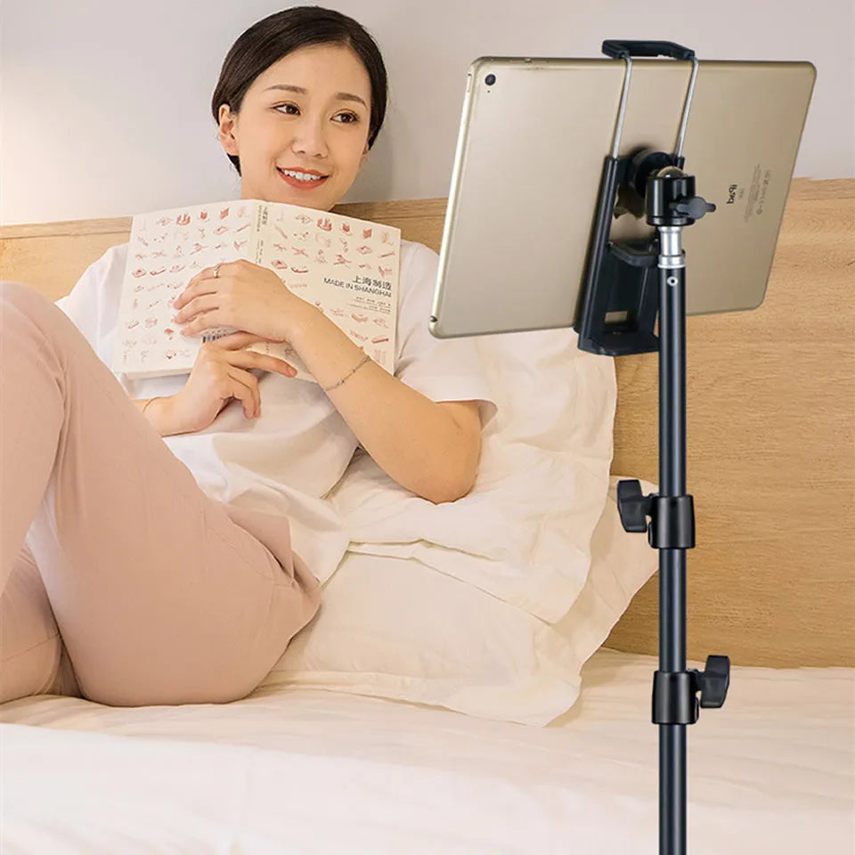 A woman is reading a book while using an adjustable aluminum tripod stand to hold a tablet.