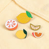 A set of enamel fruit brooches featuring lemons and oranges on a light yellow fabric background.