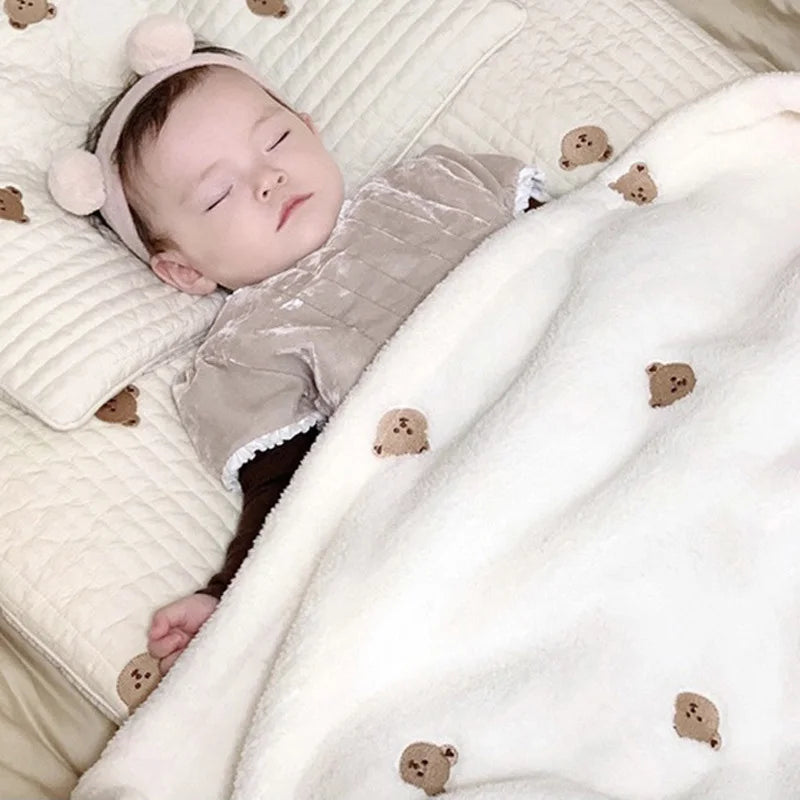 A baby is sleeping on a white coral fleece blanket decorated with small bear embroidery.
