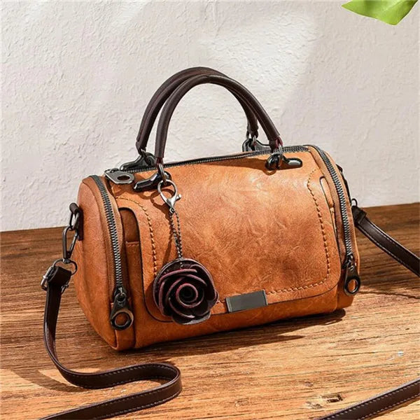 A brown leather crossbody shoulder bag with a floral pendant resting on a wooden surface.