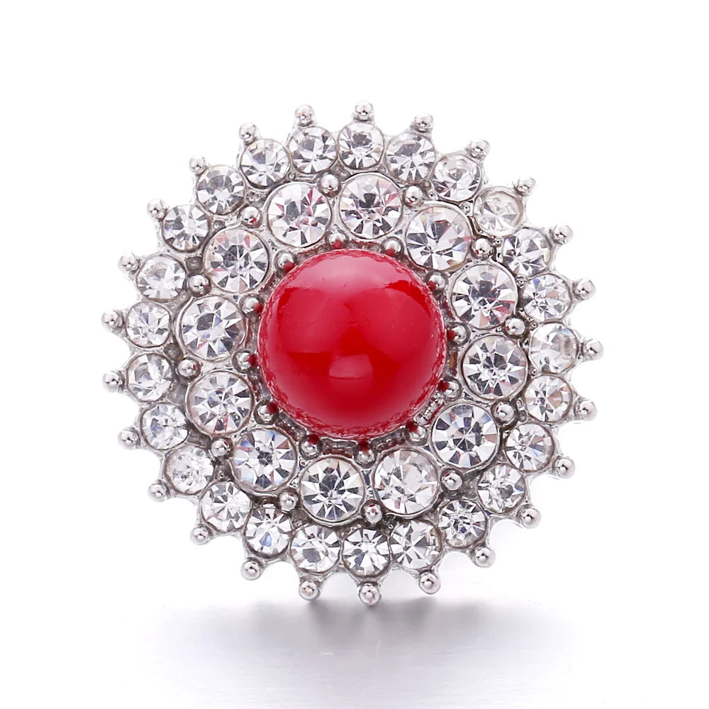 A round, brilliant silver-toned metal snap button with a central red gemstone surrounded by a halo of smaller clear stones.