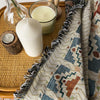 A woven throw blanket with a bohemian jacquard pattern is displayed next to candles and a glass bowl on a wooden tray.