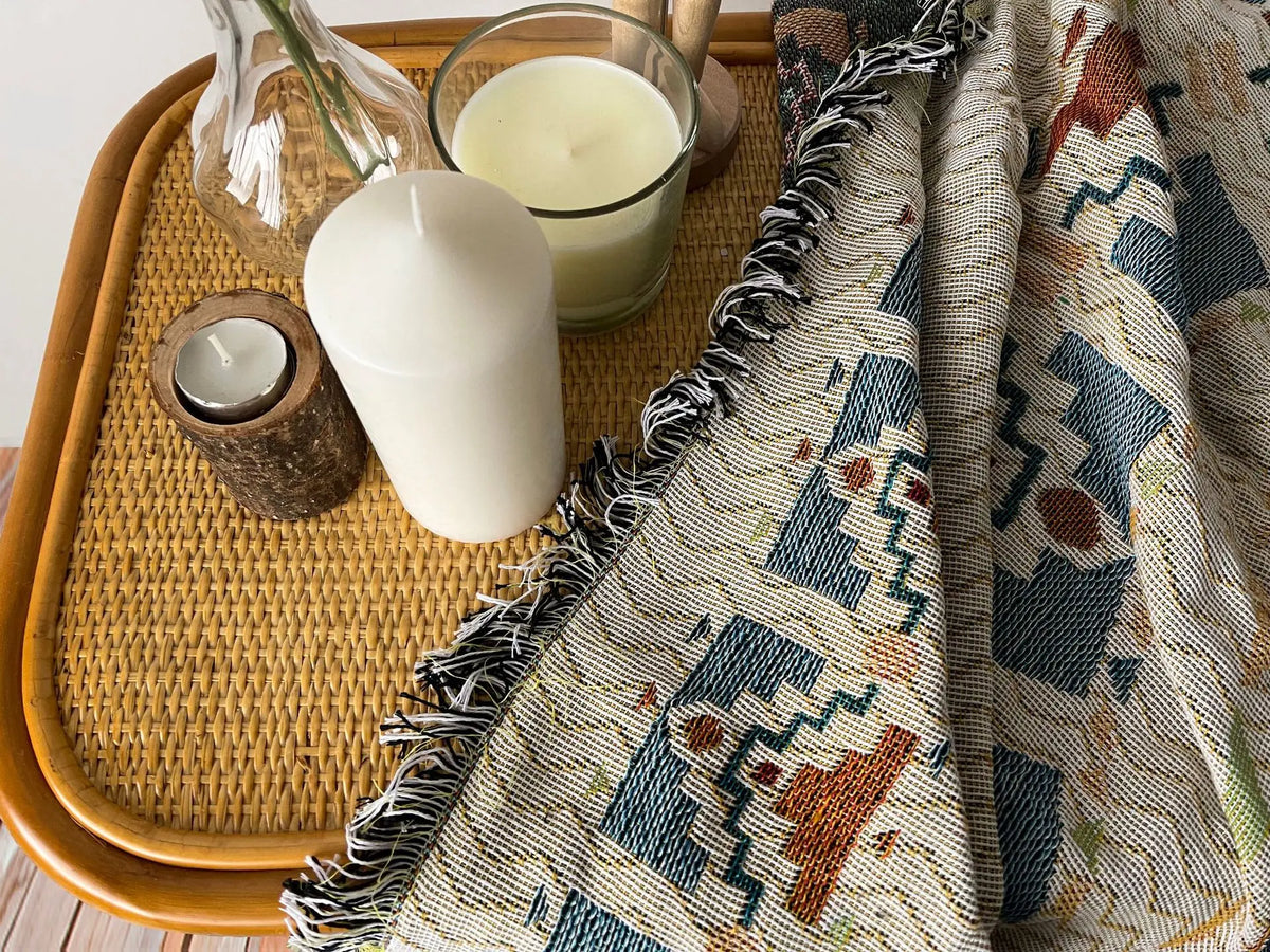 A woven throw blanket with a bohemian jacquard pattern is displayed next to candles and a glass bowl on a wooden tray.
