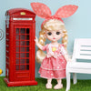 A blonde fashion doll with pink ears and a pink dress stands next to a red British telephone box.