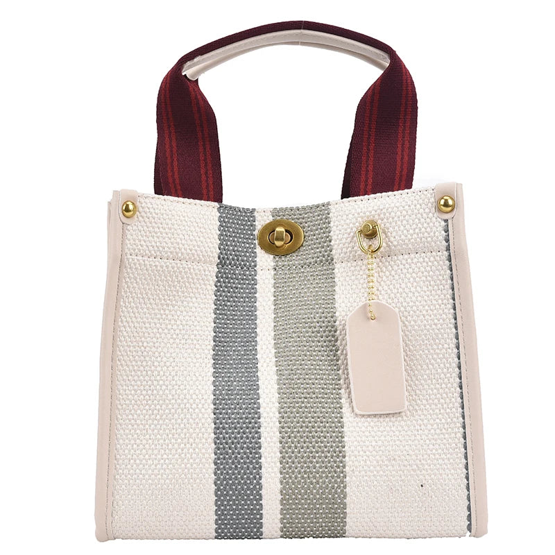 A canvas crossbody tote bag with a light beige background and vertical gray stripes, featuring a maroon strap and gold hardwa
