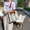 A person wearing a white t-shirt and light pants carries a beige canvas crossbody tote bag with a red strap on a city sidewal