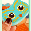 A hand holds a wooden 3D owl puzzle piece with yellow and black markings against an orange background.