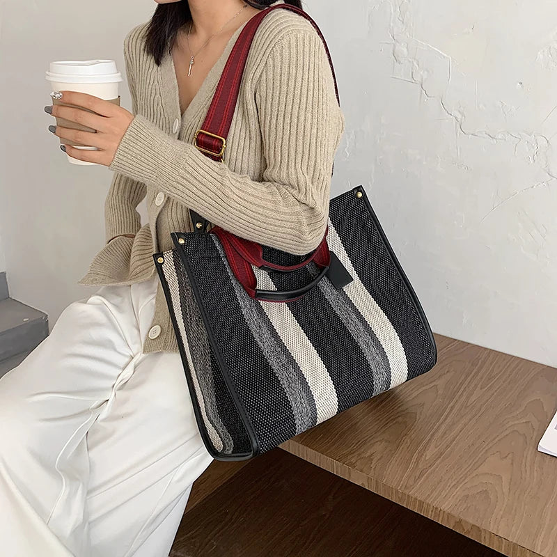 A woman holds a beige knit sweater and a white cup while resting beside a black and white striped canvas crossbody tote bag.