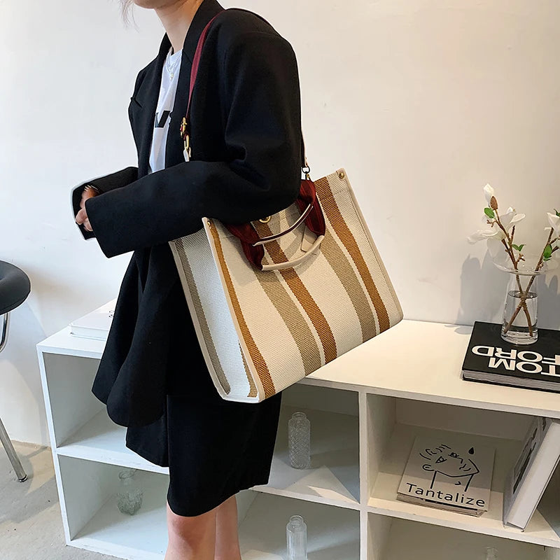 A person holds a beige and brown striped canvas crossbody tote bag against a white shelf.